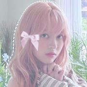 ID: Lily from Nmixx. The photo is bust upwards. She is Korean-American with pastel pink hair & bangs & a neutral expression. Her hands are grasped together under her chin. She is surrounded by green plants. There is a pink bow edited onto her hair. End ID.