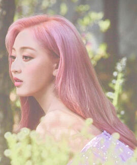 ID: Jihyo from Twice. She is in a green forest. She is a Korean woman facing the left. She has pastel pink hair and she looks wistful. The photo is edited to be muted and more pastel.